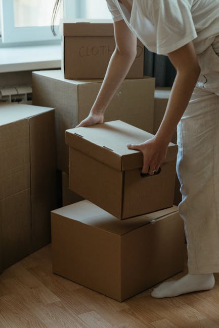 Close-up of a person wearing a dark blue T-shirt and dark trousers, positioning a large cardboard box with a caution label that reads 'CAUTION THIS SIDE UP' and features a red upward arrow. The indivi