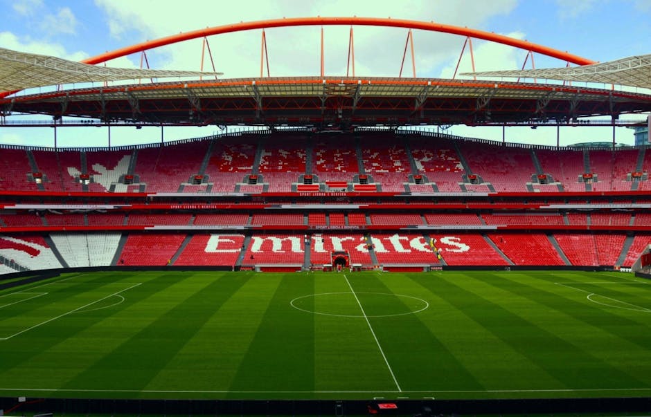 A view from the stands of Emirates Stadium showing rows of empty red plastic seats, arranged in a curved pattern facing the pitch. The seating area is clean and well-maintained, with some metal railin